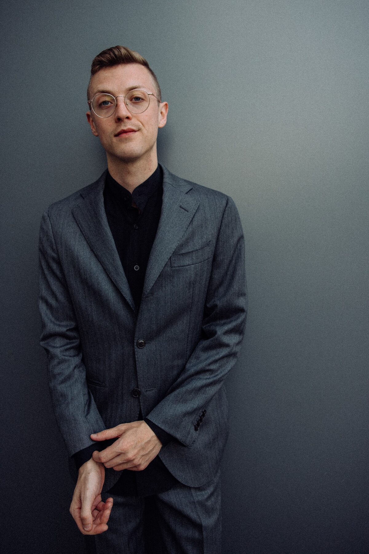Danny Killea in a grey suit and wire glasses against a slate wall